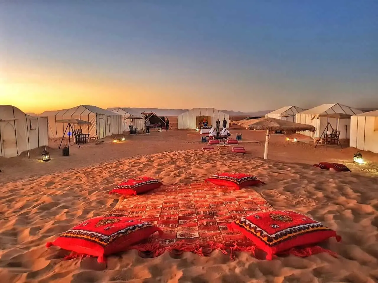 Luxury desert camp in Morocco at sunset with traditional Berber tents, red carpets, and seating areas on golden sand