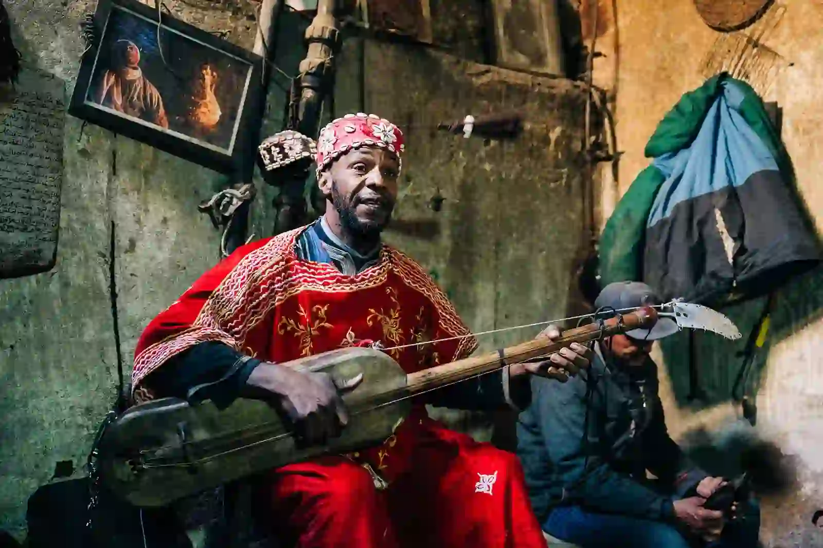 Gnawa Musician Local Encounter Gnawa musician in red attire playing guembri inside a Moroccan home.